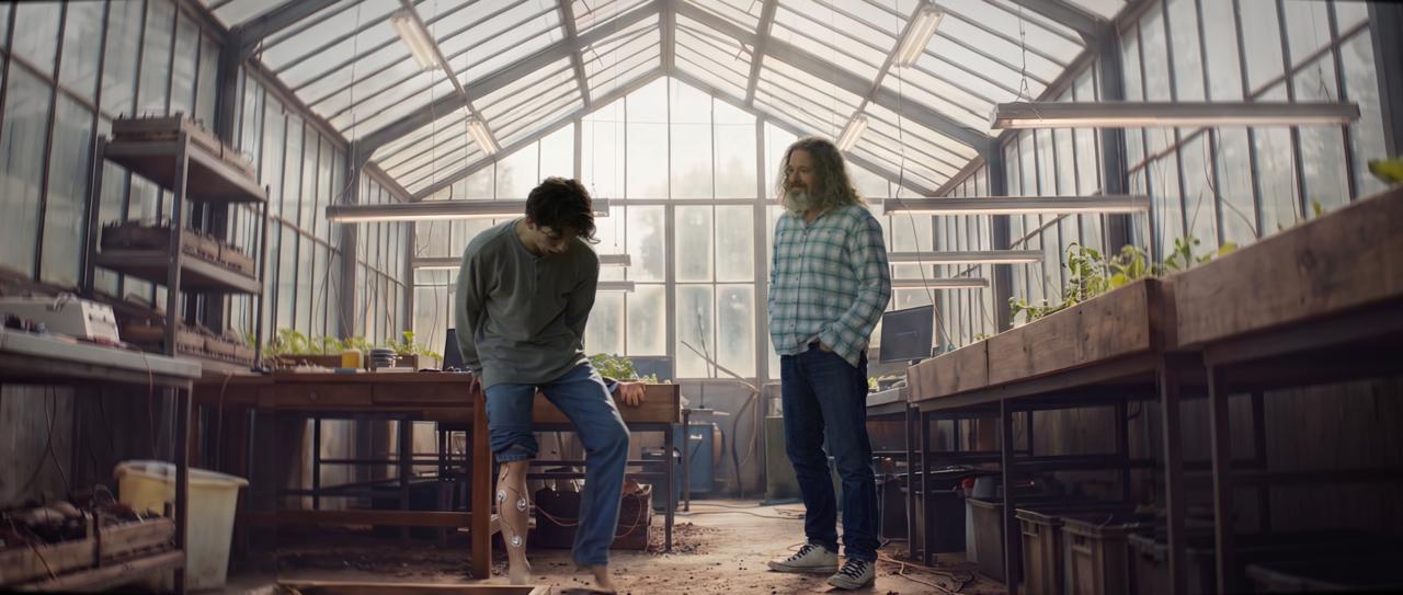 Final composite: the same two actors inside a sunlit greenhouse, electrodes on the actor's leg
