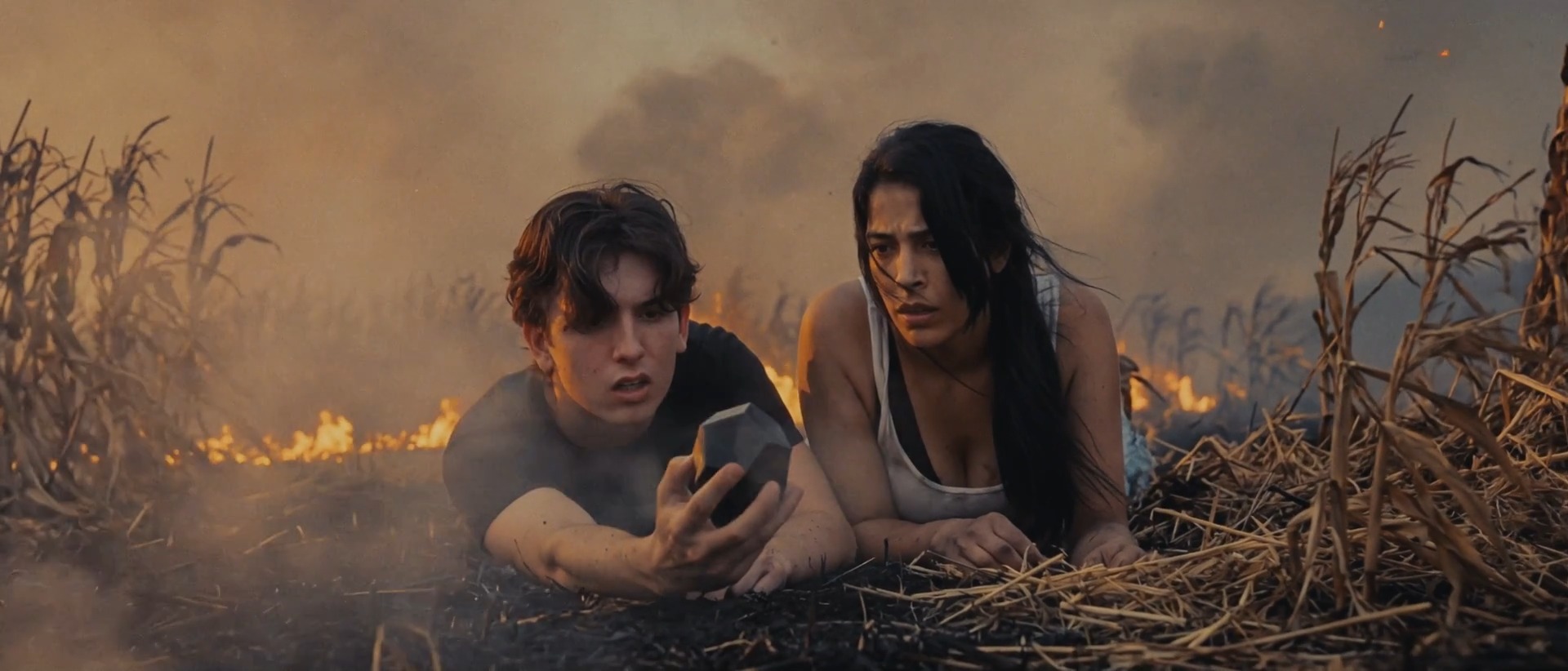 Two young people lie in a burning field; one holds a dark cube.